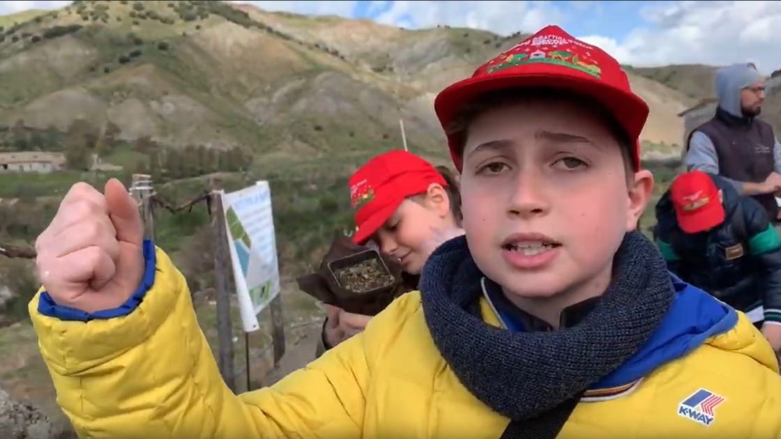 Junge mit gelber Jacke und roter Kappe steht vor Landschaft des Ätnas und spricht in die Kamera